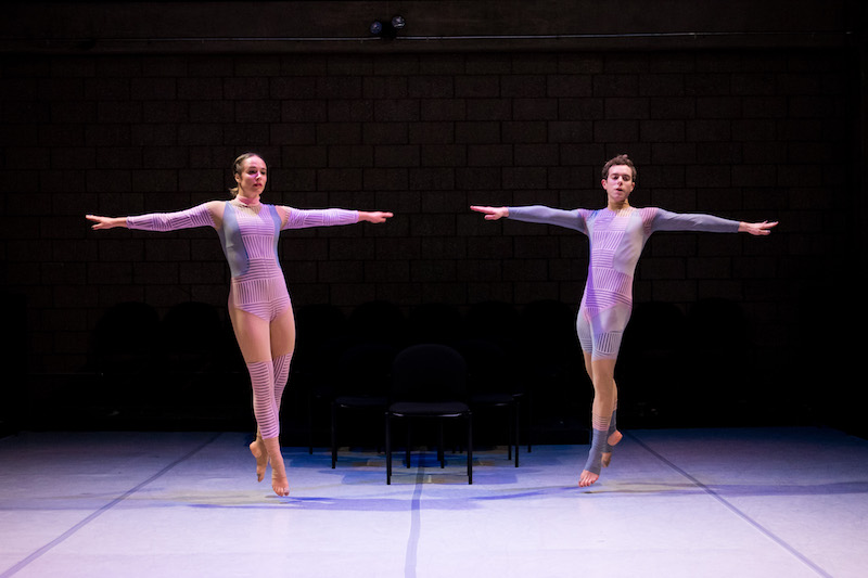 The two dancers both jump into the air towards the center of the stage in a symmetrical configuration with deadpan stares